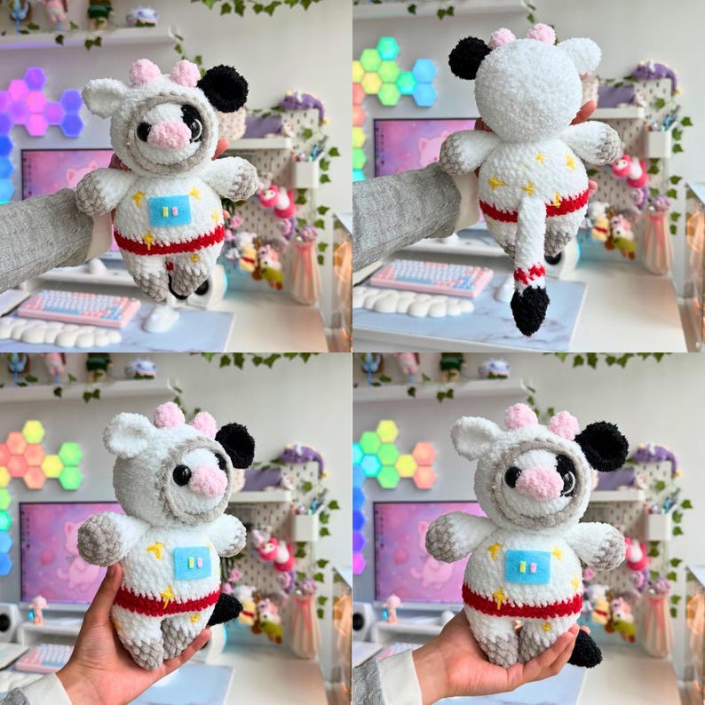 Op de afbeelding: Een handgemaakte gehaakte koeienknuffel in een ruimtepakontwerp. De knuffel is voornamelijk wit met zwarte en roze accenten, een rode streep en een blauw vierkant met gele sterren. De koe heeft een capuchon en een staart.