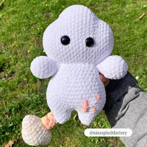 Op de afbeelding: Een handgemaakt wit gehaakt pluche speelgoed met zwarte knoopogen, kleine armen en een kleurrijke staart. Het pluche heeft een wolkvorm en is versierd met roze stervormige accenten. Het speelgoed wordt tegen een achtergrond van groen gras gehouden.