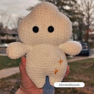 Op de afbeelding: Een cr&egrave;mekleurige, handgemaakte gehaakte knuffel in de vorm van een wolk of paddenstoel. Het heeft twee zwarte knoopogen, kleine armen en oranje kruissteekdetails. De knuffel wordt in een hand gehouden.