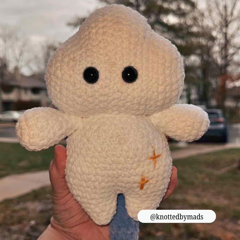 Op de afbeelding: Een cr&egrave;mekleurige, handgemaakte gehaakte knuffel in de vorm van een wolk of paddenstoel. Het heeft twee zwarte knoopogen, kleine armen en oranje kruissteekdetails. De knuffel wordt in een hand gehouden.