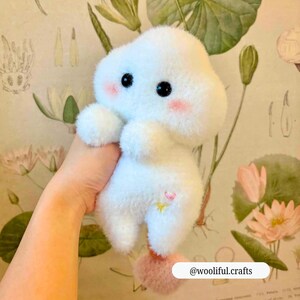 Op de afbeelding: Een zacht, wit, wolk-vormig pluche speelgoed met zwarte ogen en roze wangen. Het heeft een klein roze hart en gele ster. Een roze pompon is aan de onderkant bevestigd. De tekst "@wooliful.crafts" is zichtbaar.