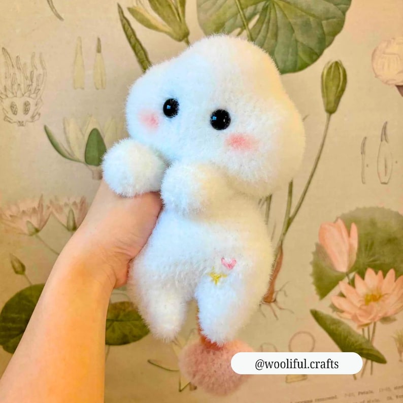 Op de afbeelding: Een zacht, wit, wolk-vormig pluche speelgoed met zwarte ogen en roze wangen. Het heeft een klein roze hart en gele ster. Een roze pompon is aan de onderkant bevestigd. De tekst "@wooliful.crafts" is zichtbaar.