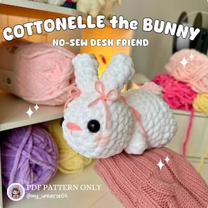 May include: A white crocheted bunny with pink accents, including a bow and blush. The text "Cottonelle the Bunny" and "No-Sew Desk Friend" are displayed. The image also shows various yarn colors.