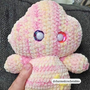 Op de afbeelding: Een handgemaakt gehaakt knuffel in roze, geel en wit. Het speelgoed heeft grote, kleurrijke, glinsterende ogen en kleine armen. De tekst "@charmedcrochetables" is rechtsonder zichtbaar.