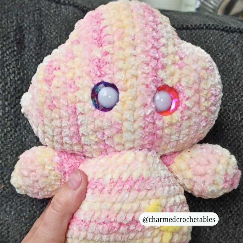 Op de afbeelding: Een handgemaakt gehaakt knuffel in roze, geel en wit. Het speelgoed heeft grote, kleurrijke, glinsterende ogen en kleine armen. De tekst "@charmedcrochetables" is rechtsonder zichtbaar.