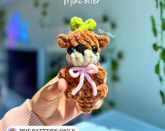 May include: A brown crocheted mini otter plush with a light green leaf on its head, a cream-colored snout, black eyes, and a light pink ribbon tied around its neck. The text "Mini otter" and "No-sew!" appear above, with "PDF PATTERN ONLY" below.
