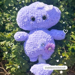 Op de afbeelding: Een handgemaakt lavendelkleurig pluche speelgoed met grote zwarte ogen, een witte strik en twee decoratieve bloemaccenten. Het pluche is omgeven door groene bladeren en heeft de tekst "@adelinecrochetcraft" onderaan.