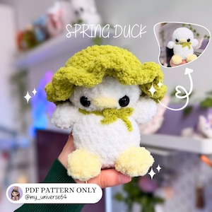 May include: A handmade white and yellow crochet duck with a green hat and bow. The text "SPRING DUCK" is above the duck. The image also includes a smaller image of the duck and the text "PDF PATTERN ONLY @my_universe64".