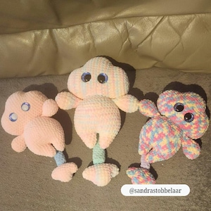 Op de afbeelding: Drie handgemaakte gehaakte speelgoed in de vorm van een wolk. Het speelgoed is in pastelkleuren, met grote, blauwe wiebelogen. E&eacute;n is roze, &eacute;&eacute;n is veelkleurig en &eacute;&eacute;n is een mix van roze en pastelkleuren. Het speelgoed heeft armen en benen.