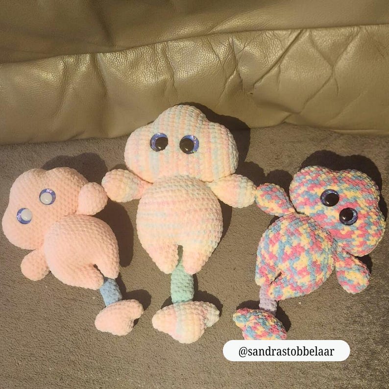 Op de afbeelding: Drie handgemaakte gehaakte speelgoed in de vorm van een wolk. Het speelgoed is in pastelkleuren, met grote, blauwe wiebelogen. E&eacute;n is roze, &eacute;&eacute;n is veelkleurig en &eacute;&eacute;n is een mix van roze en pastelkleuren. Het speelgoed heeft armen en benen.