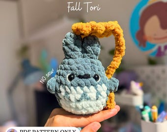 May include: A blue and white crocheted plush toy shaped like a creature with two ears, small arms, and a textured mustard yellow leaf accessory. The text Fall Tori, NO sew!, and PDF PATTERN ONLY @my_universe64 are visible on the image.