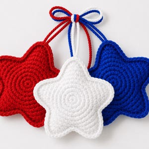 May include: Three star-shaped ornaments crocheted in solid red, white, and blue yarn. Each star features a circular stitch pattern and is attached to a matching colored cord. The three cords are gathered together at the top and tied in a bow.