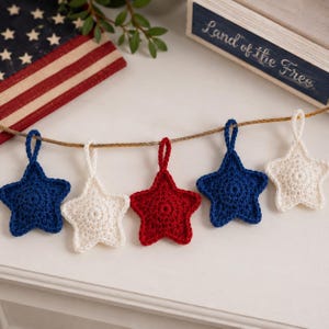 May include: A garland featuring five crocheted star ornaments in red, white, and blue, suspended from a thin brown twine. The stars are arranged in a repeating pattern of blue, white, red, blue, and white, each with a small loop for hanging.