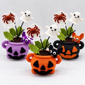 May include: Three crocheted Halloween pots in purple, orange, and black. Each pot features a jack-o'-lantern face and contains crocheted green leaves, white spiderwebs with brown spiders, and small figures including ghosts and bats.