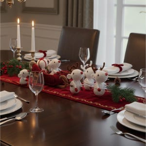 May include: A festive dining table setting with a red runner, white plates, and wine glasses. Crochet reindeer and a sleigh are the centerpiece, with lit candles and greenery adding to the holiday theme. The table is set for a special occasion.
