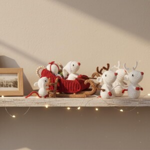 May include: Crocheted Christmas decorations featuring a red sleigh pulled by white reindeer with brown antlers. The sleigh carries white mice and a wrapped gift. The decorations are displayed on a white shelf with string lights.