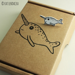 May include: A brown cardboard box with a black and white illustration of a narwhal on the front. A smaller, white narwhal sticker is placed on the top of the box.