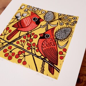 May include: A yellow background with two red cardinals perched on branches. The cardinals are stylized with black outlines and red and black details. The background is filled with black and white berries and leaves.