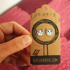May include: A pair of white cat face stud earrings on a brown card with a black circle and a small red bird illustration. The card also has the text "KATLENDACKA.COM".
