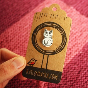 May include: A brown paper tag with a white cat illustration inside a circle. The tag has the text "KATLENDACKA.COM" printed on the bottom. A small red bird illustration is on the left side of the tag.