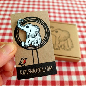 May include: A white elephant pin with black outlines and red cheeks, sitting on a tree branch. The pin is on a brown card with the text "katlendacka.com".