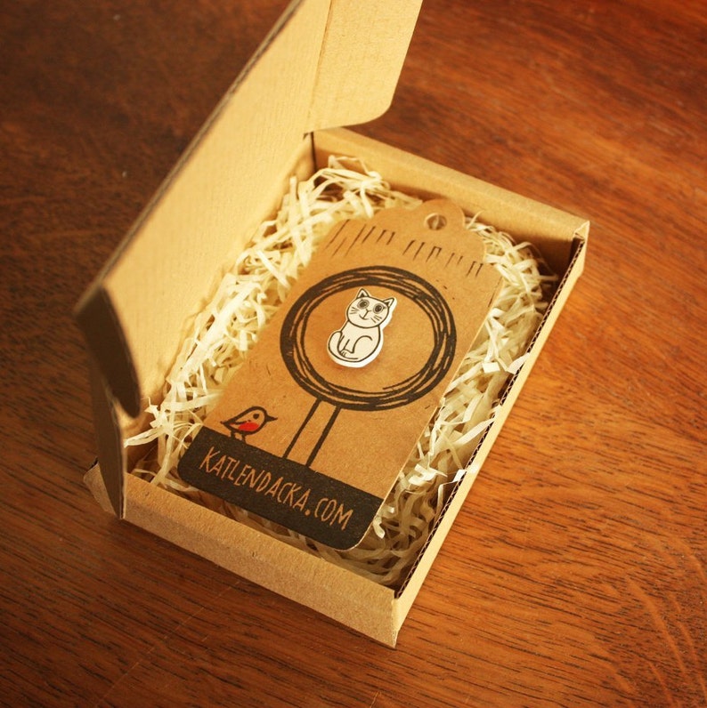 May include: A white cat pin in a cardboard box with brown packing paper. The pin is on a brown tag with a black circle around it and a red bird illustration. The tag says "KATLENDACKA.COM".