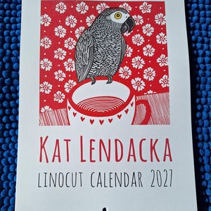 May include: A spiral-bound wall calendar titled Kat Lendacka Linocut Calendar 2027. The cover features a linocut print of a grey parrot perched on a white mug with red heart accents, set against a red background with a white floral pattern.