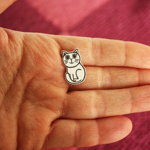 May include: A white paper cutout of a cartoon cat with black eyes and a black outline. The cat is sitting with its paws tucked under its body.