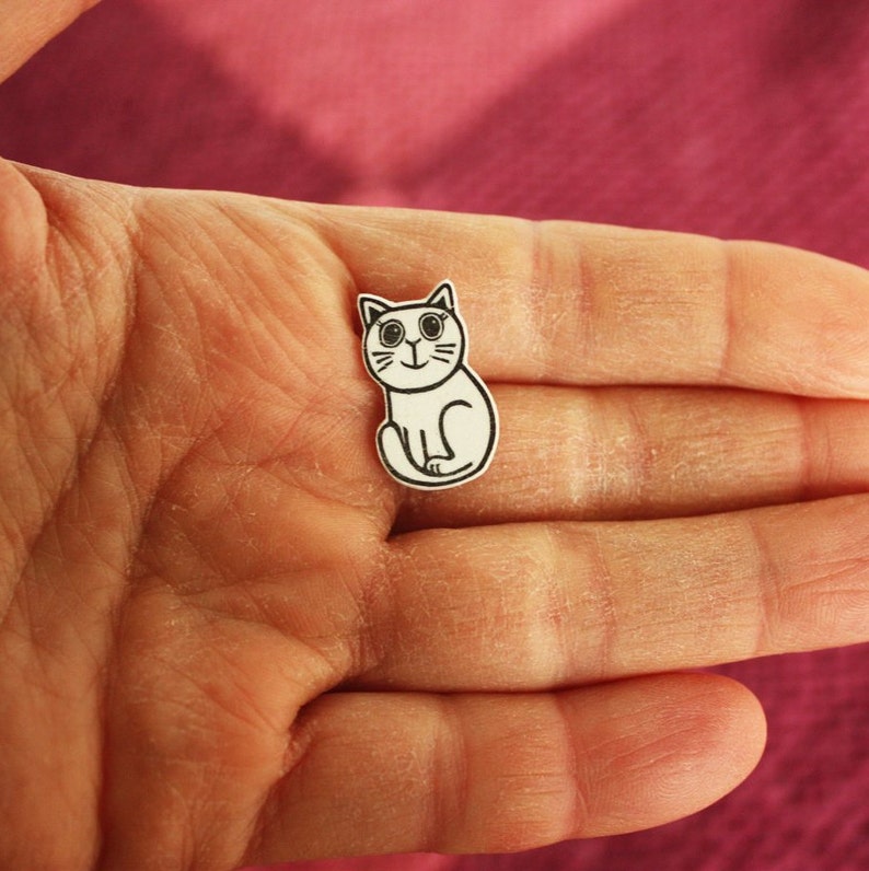 May include: A white paper cutout of a cartoon cat with black eyes and a black outline. The cat is sitting with its paws tucked under its body.