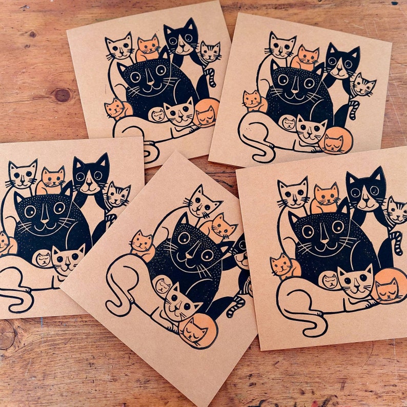 May include: A collection of square cards with a black and orange cat illustration. The cats are depicted in a whimsical style, with a large black cat surrounded by smaller orange and black cats. The cards are printed on a kraft paper background.
