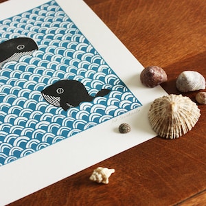 May include: A blue and white print of two whales swimming in the ocean. The print is on white paper and is surrounded by seashells.