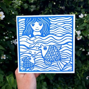 May include: A square, blue and white illustration of a woman in water with two fish. The woman has long hair and is smiling. The fish are swimming in wavy lines. The artwork is held in front of a green leafy background.