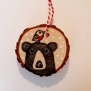 May include: A round wooden ornament with a hand-painted bear and bird design. The bear is black with a white mouth and eyes, and the bird is red and black. The ornament has a red and white string for hanging.