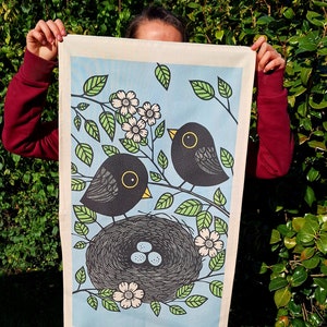 May include: A tea towel with a blue background featuring a blackbird family. The blackbirds are perched on branches with white flowers. The blackbird on the left is sitting on a nest with three white eggs. The tea towel has a white border.