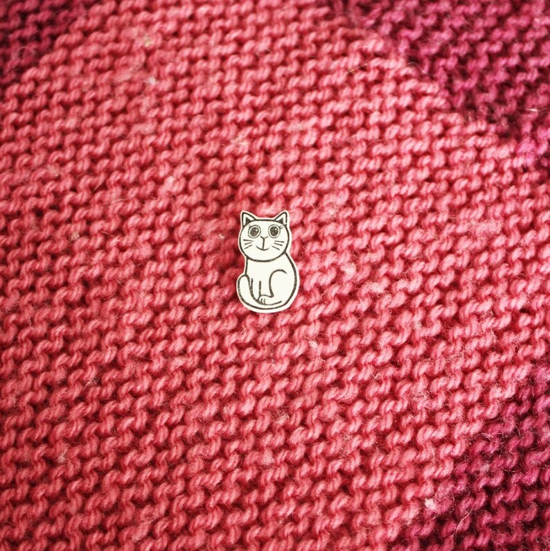 May include: A white cat pin with black eyes and a pink nose, sitting on a red knitted fabric.