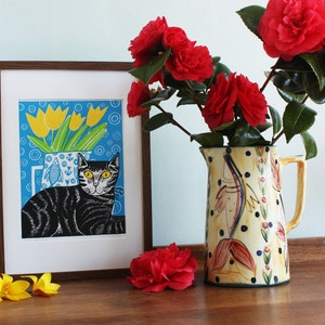 May include: A framed print of a black and white cat with yellow eyes, sitting in front of a blue vase with yellow tulips. The vase is on a blue background with white circles. The print is in a brown frame and is sitting on a wooden table with a red camellia flower in front of it.