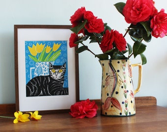 May include: A framed print of a black and white cat with yellow eyes, sitting in front of a blue vase with yellow tulips. The vase is on a blue background with white circles. The print is in a brown frame and is sitting on a wooden table with a red camellia flower in front of it.