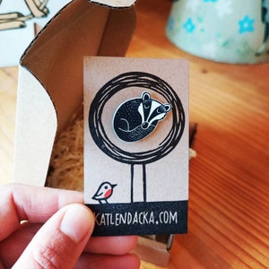 May include: A black and white enamel pin featuring a badger curled up asleep inside a circle. The pin is on a brown card with a small bird illustration and the text "katlendacka.com".
