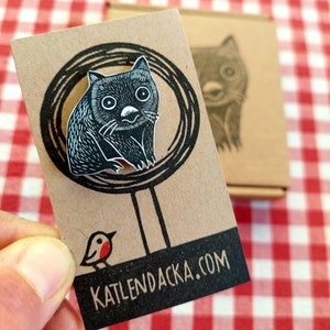 May include: A black and white illustration of a wombat on a brown card with a circle around it. The card has the text "katlendacka.com" printed on it.