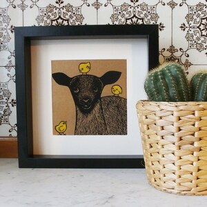 May include: A black and white illustration of a black lamb with three yellow chicks on its head and one chick standing in front of it. The illustration is framed in a black frame and is displayed on a white surface.