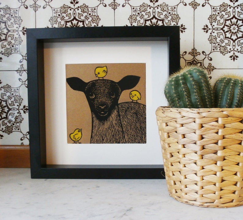 May include: A black and white illustration of a black lamb with three yellow chicks on its head and one chick standing in front of it. The illustration is framed in a black frame and is displayed on a white surface.