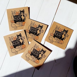 May include: A collection of square, kraft paper cards featuring a black and orange cat illustration. The design includes a large cat surrounded by smaller cats. The cards are arranged on a white wooden surface.