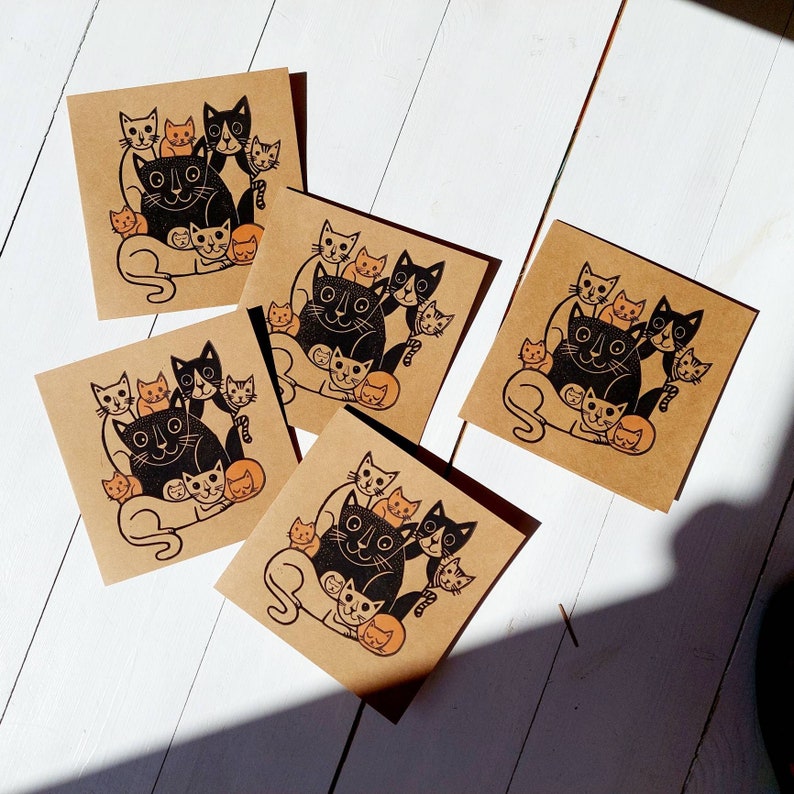 May include: A collection of square, kraft paper cards featuring a black and orange cat illustration. The design includes a large cat surrounded by smaller cats. The cards are arranged on a white wooden surface.