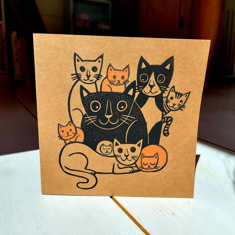 May include: A square greeting card with a brown background features a black and orange cat illustration. The illustration depicts several cats of varying sizes, with the largest cat in the center. The cats have simple, stylized features.