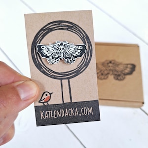May include: Black and white enamel pin featuring a moth with a skull design. The pin is displayed on a brown card with the website "KATLENDACKA.COM" printed on it.
