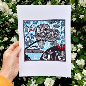 May include: A black and white linocut print of two owls perched on a branch with red berries. The owls are surrounded by swirling blue lines and a small house is visible in the background. The print is titled "The Owls" and signed by the artist.