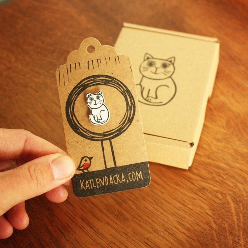 May include: A brown paper tag with a white cat illustration, a red bird illustration, and the text "KATLENDACKA.COM". The tag is attached to a brown cardboard box.
