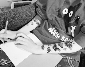 May include: Black and white image of a person wearing a dark red sweatshirt with a cat graphic and the text "NO SUN". The person has butterfly and skull temporary tattoos on their arms and is holding a pencil and a notebook.