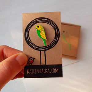 May include: A green and yellow parakeet pin on a brown card with a black circle and the text "KATLENDACKA.COM".
