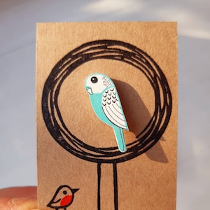 May include: A light blue and white wooden bird pin with a black outline. The pin is on a brown card with a black circle around it.